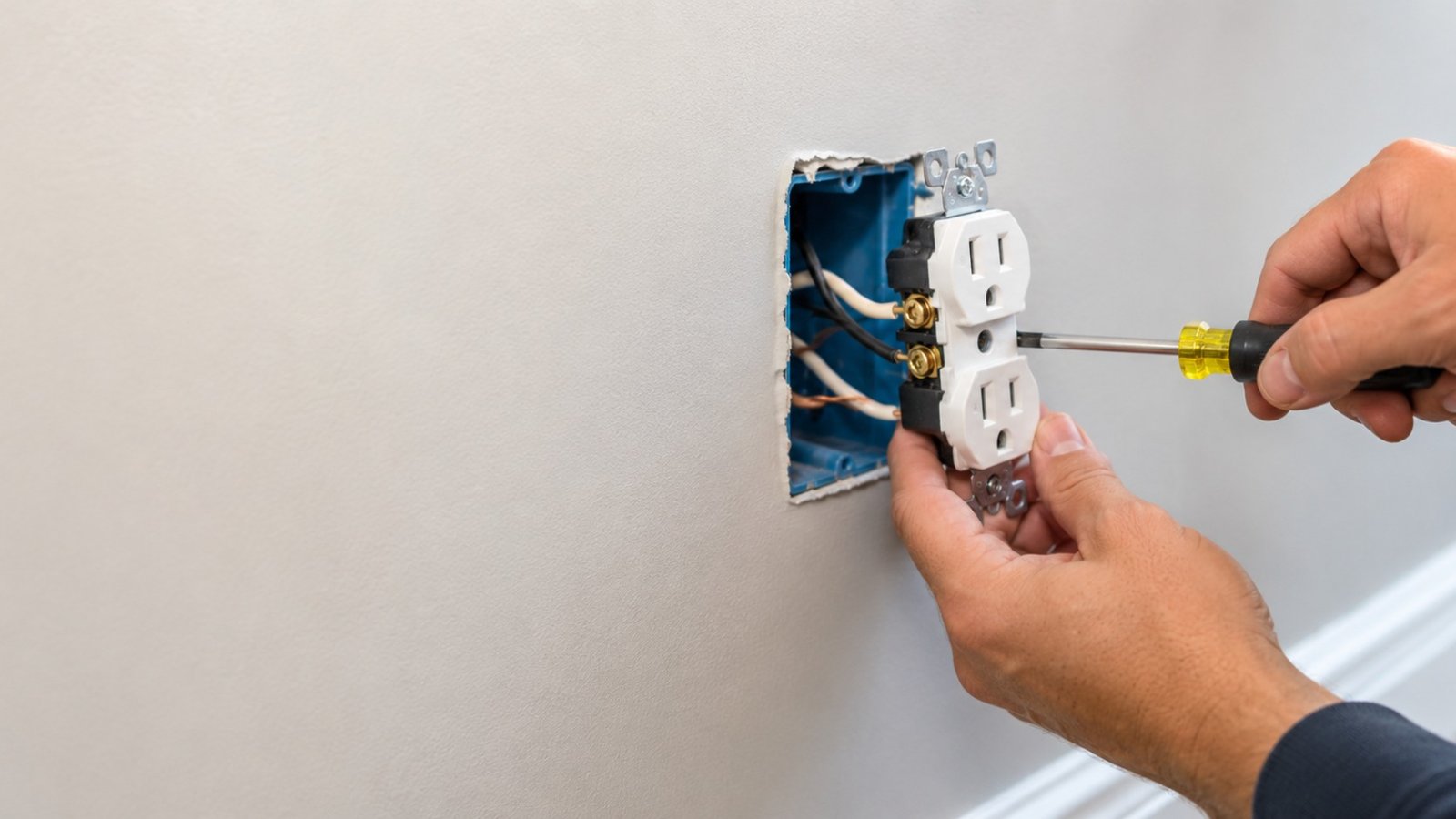 Electrician's hands installing a grounded outlet with wiring visible in an open wall box.